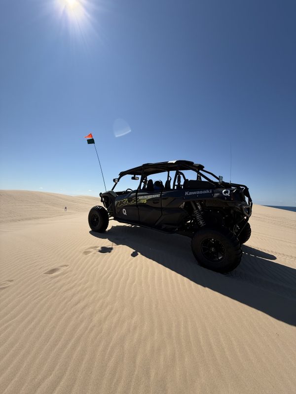 Off-road vehicle on sand dunes under a clear blue sky with sun.