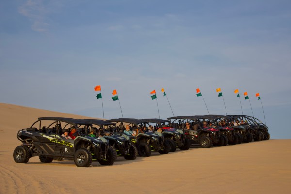 Row of dune buggies with flags parked on sandy desert hill under blue sky.