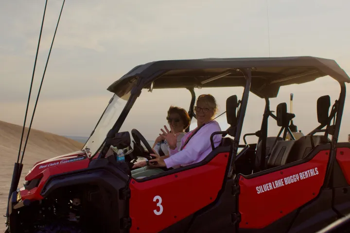 Two people waving from a red dune buggy in a desert landscape.