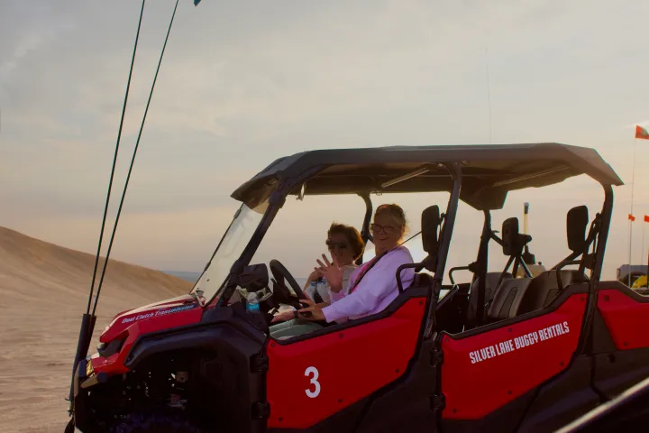 Two people waving from a red dune buggy in a desert landscape.