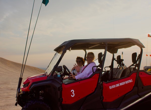 Two people waving from a red dune buggy in a desert landscape.
