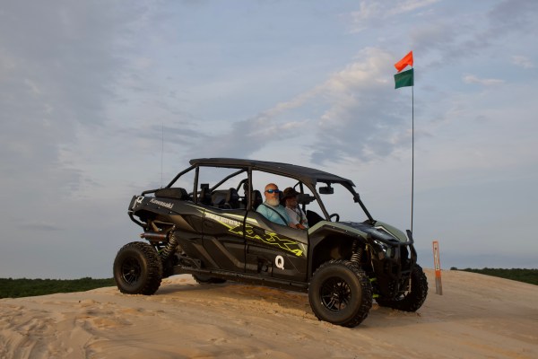 An ATV with two people on a sand dune, under a cloudy sky. An orange and green flag is attached to the ATV.