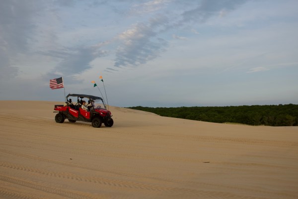 Off-road vehicle with flags driving on sand dunes under a cloudy sky.