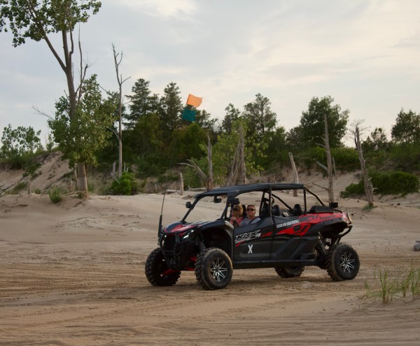 ATV with passengers driving on sandy terrain with trees in the background.