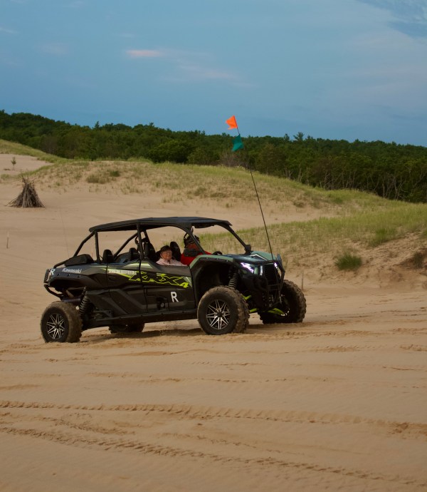 Off-road vehicle with flags driving on sandy dunes under a blue sky.