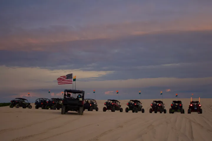 Off-road vehicles with flags on sandy terrain under a cloudy sky at sunset.