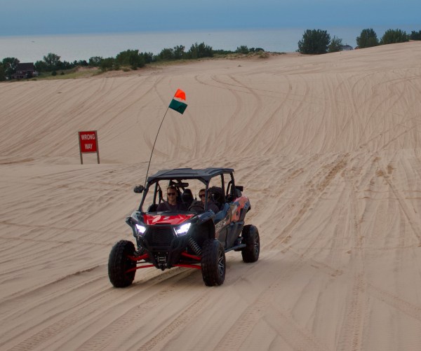 ATV with two people on sand dunes, red sign saying 'Wrong Way' in background, trees and sky visible.