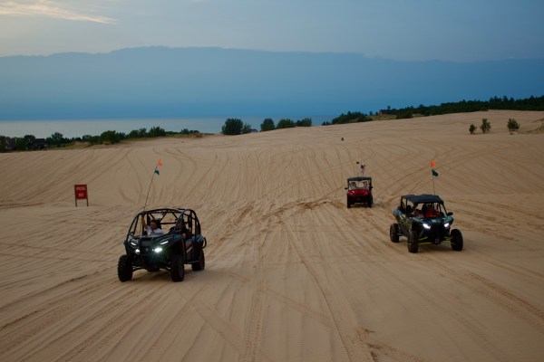 Three dune buggies driving on sand dunes with a lake and trees in the background.