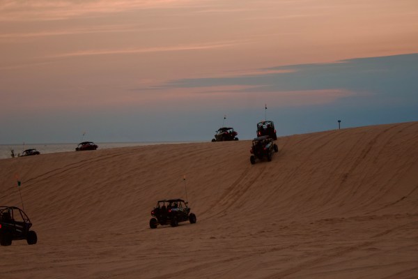 ATVs driving on sand dunes under a colorful sunset sky.