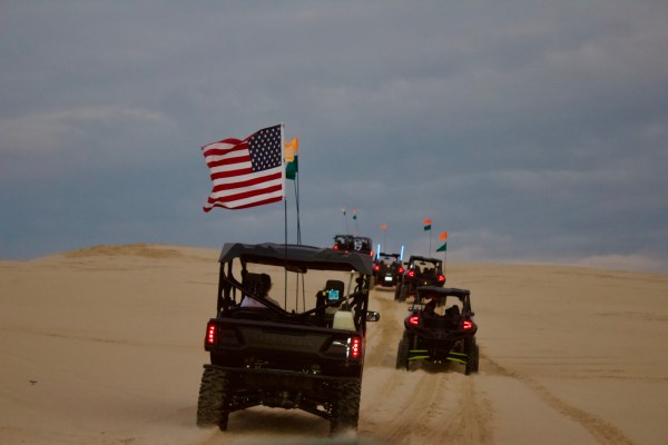 ATVs with flags driving up sandy hill under cloudy sky.