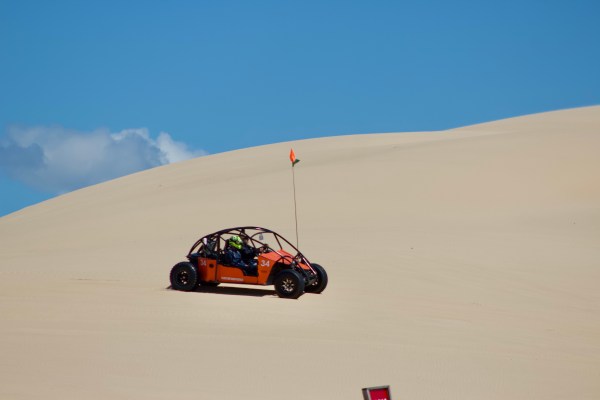 Orange dune buggy driving on sand dunes under a clear blue sky.