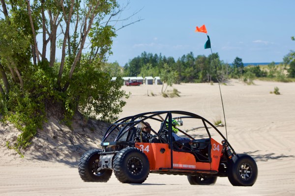 Orange dune buggy with a flag driving on sandy terrain near trees.