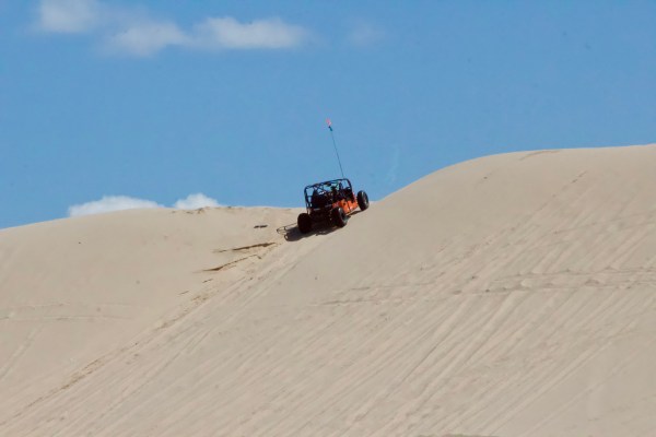 A dune buggy driving up a large sand dune under a clear blue sky.