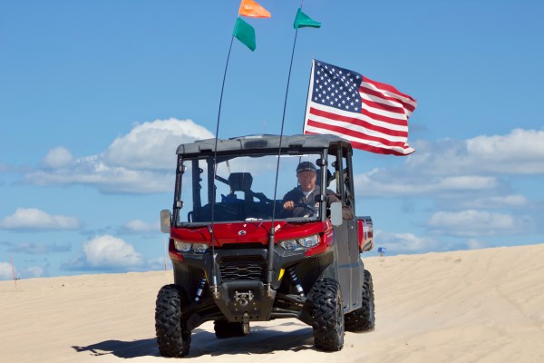 Red ATV with American flag on sand dune under blue sky with clouds.
