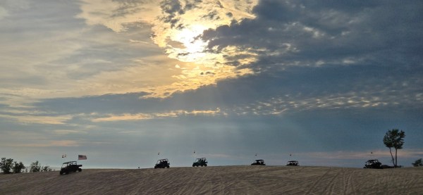 Silhouetted off-road vehicles on sand dune under cloudy sky with sun breaking through.
